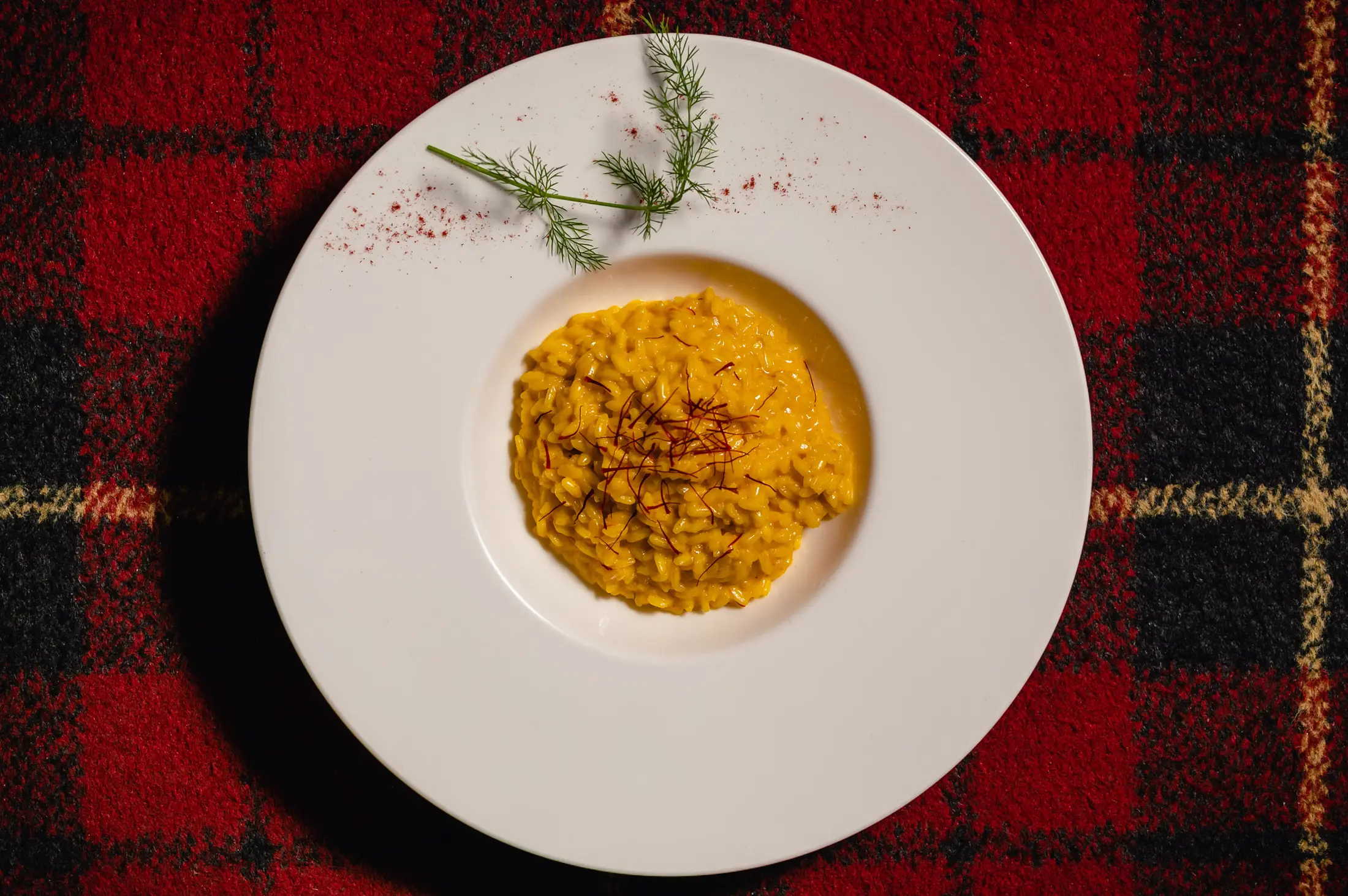 Il risotto giallo alla milanese coi pistilli di zafferano, in un piatto bianco sul tartan rosso, visto dall’alto