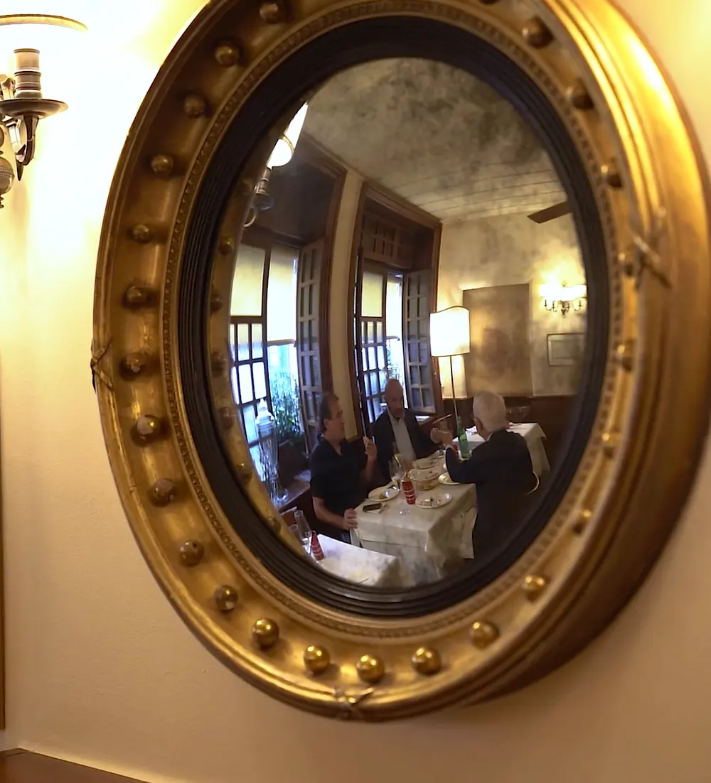 Lo specchio a occhio di bue, con la cornice dorata, che riflette la sala